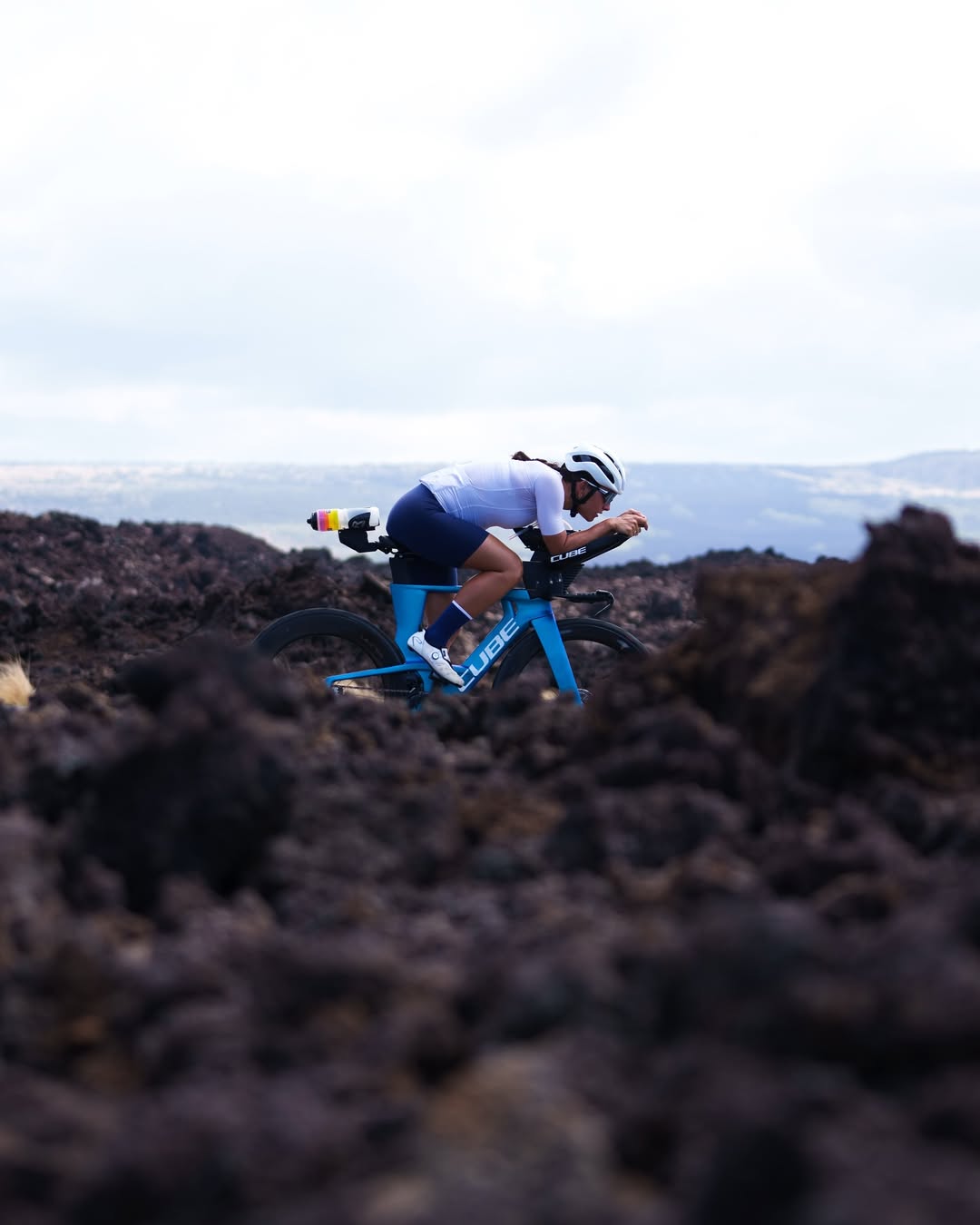 Triathlete riding a blue Cube triathlon bike through a volcanic landscape.