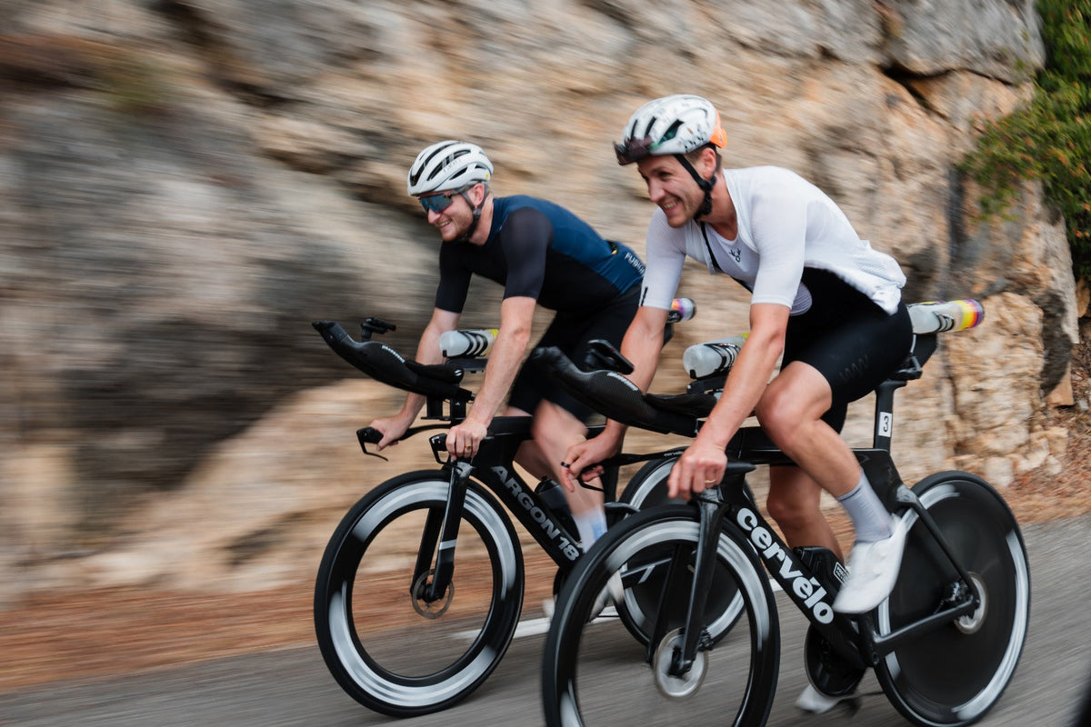 Two male cyclists riding aerodynamic triathlon bikes on a paved road with motion blur.