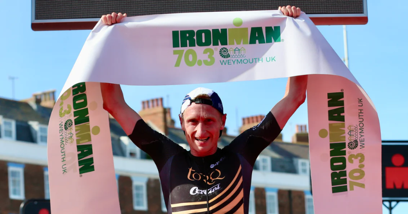 Male triathlete celebrating victory holding an Ironman 70.3 Weymouth UK finisher banner.