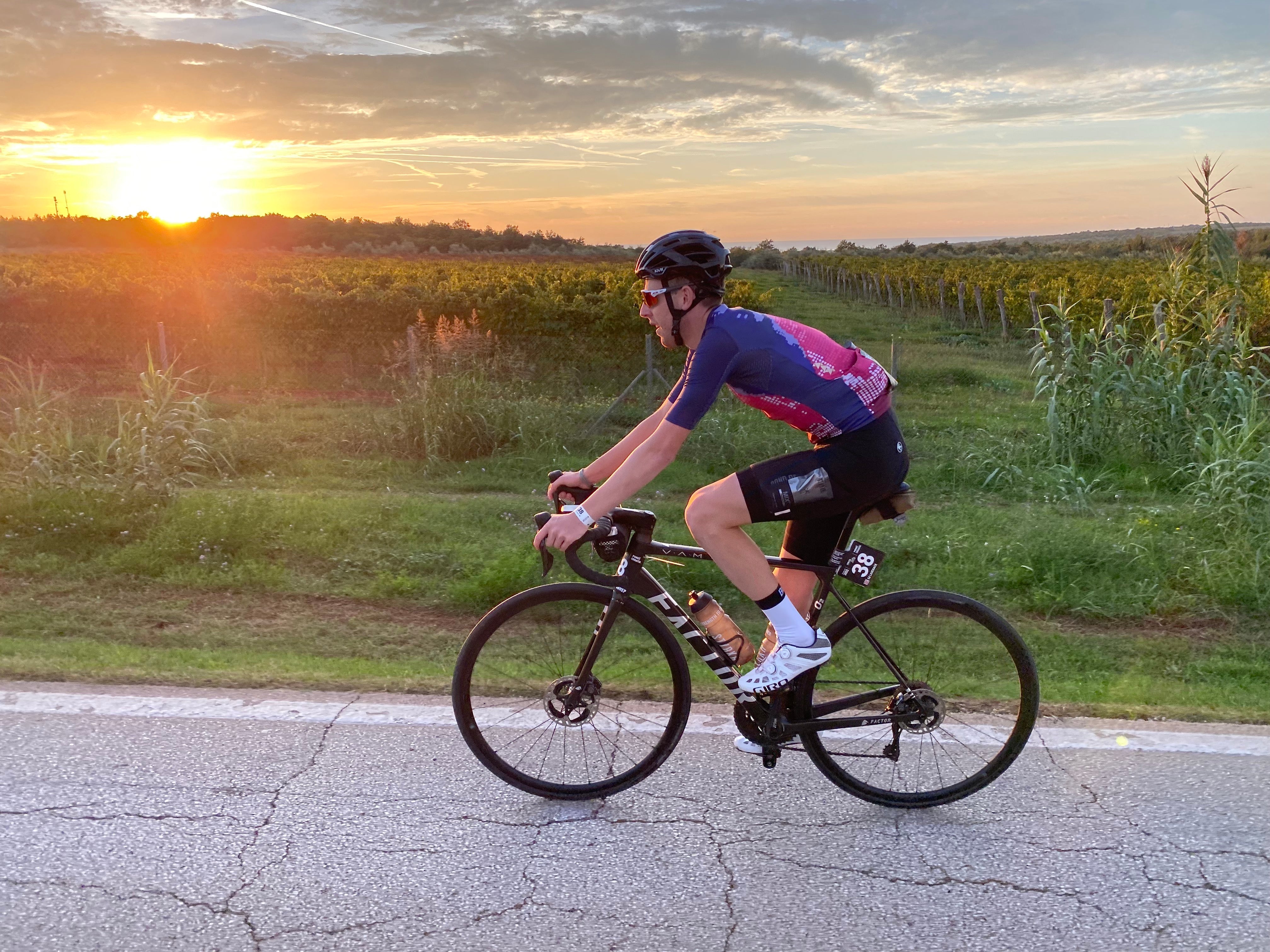 Male cyclist riding a road bike on a paved road during a sunset in a rural landscape.