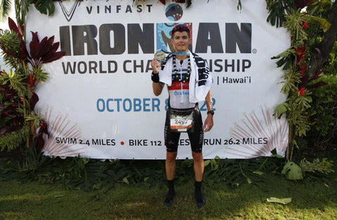 Male triathlete holding a finisher medal in front of an Ironman World Championship banner.