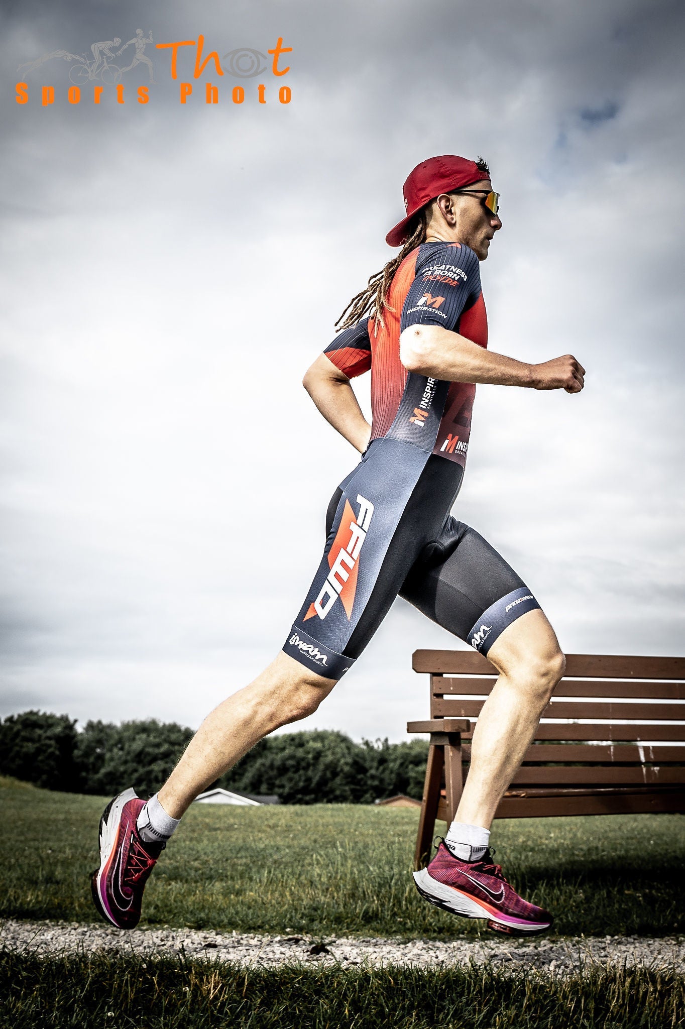 Male triathlete in red and grey racing kit running on a grass path outdoors.