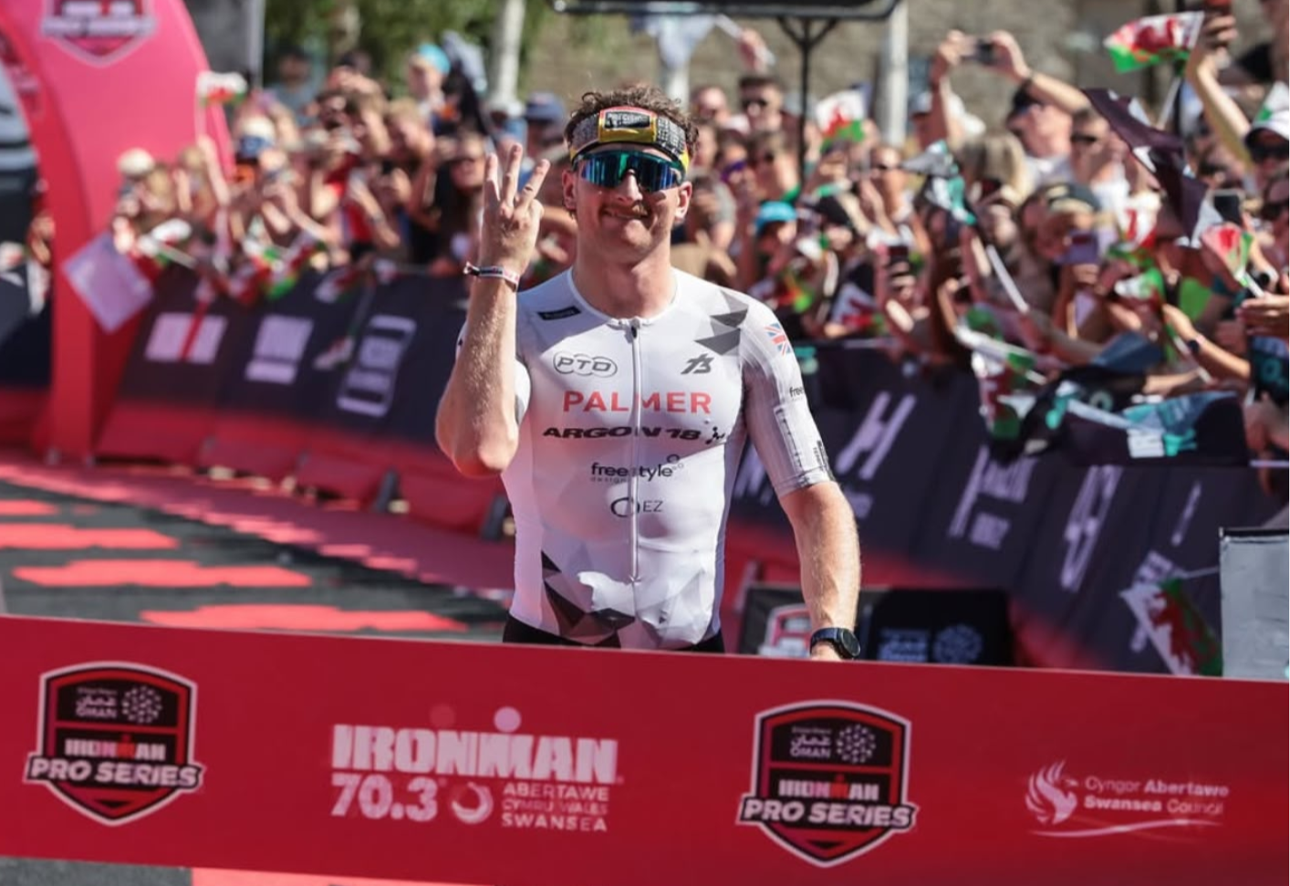 Triathlete celebrating at the Ironman 70.3 Pro Series finish line wearing white cycling jersey.